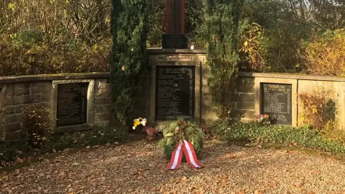 Ein steinernes Kreuzdenkmal, umgeben von Bäumen und Sträuchern, mit einem Kranz an seinem Sockel.