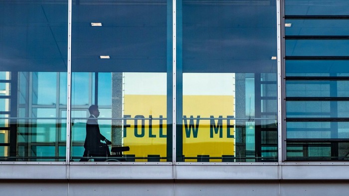 Eine Person geht hinter einer Glasfassade entlang, während im Hintergrund ein gelbes Schild mit der Aufschrift „FOLLOW ME“ durch die Fenster sichtbar ist.