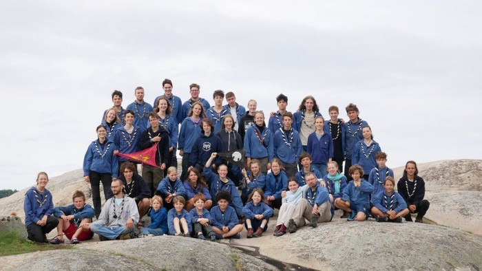 Eine große Gruppe von Menschen in überwiegend blauer Kleidung sitzt und steht gemeinsam draußen auf felsigem Gelände und blickt in die Kamera