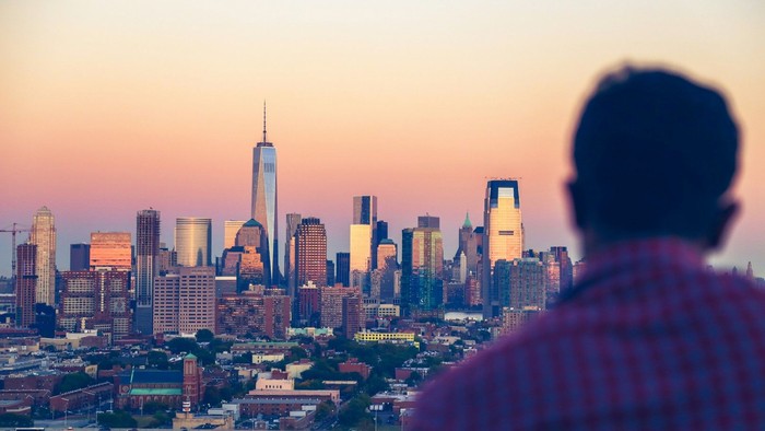 Eine einzelne Person blickt vom Stadtrand aus auf die Skyline einer großen Stadt im warmen Licht des Sonnenuntergangs.