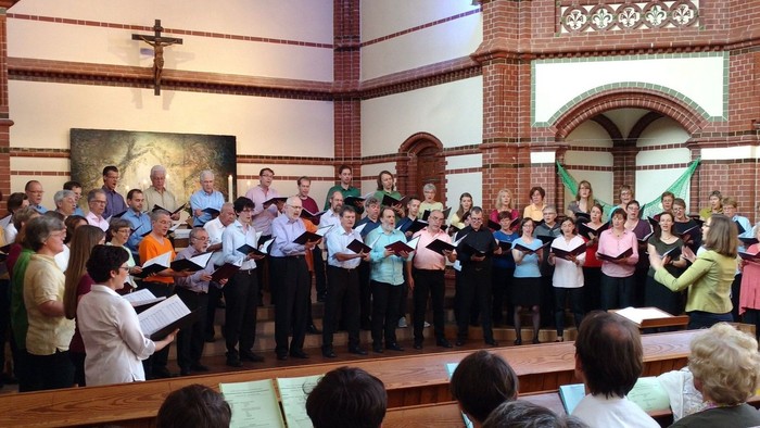 Ein großer Chor tritt in einer Kirche auf, dirigiert von einem Dirigenten, während Zuschauer im Hintergrund sitzen.