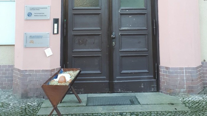 Das Bild zeigt einen pinken Hauseingang mit zwei schwarzen Türen und einem kleinen Tisch mit Informationsmaterialien.