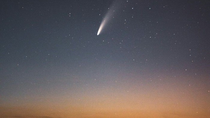 Ein Komet zieht bei Dämmerung über einem beleuchteten Ort am Himmel