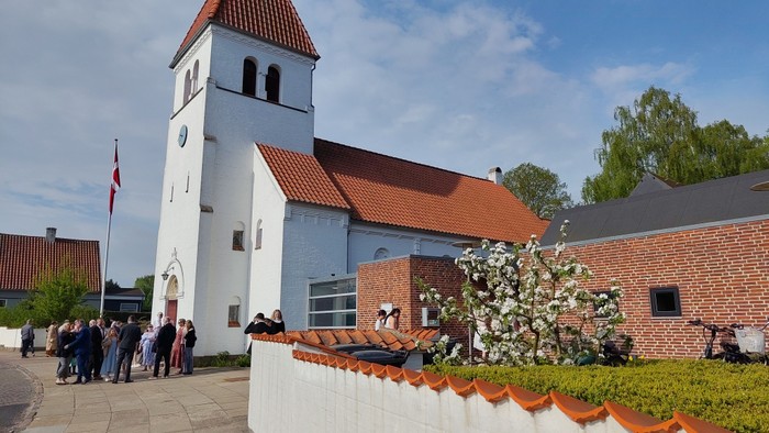 En gruppe samles uden for en hvid kirke med et højt klokketårn på en solskinsdag.
