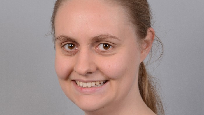 Junges Mädchen mit braunen Haaren, die zu einem Pferdeschwanz gebunden sind, lächelt in die Kamera. Sie trägt eine schwarze Jacke über einem weißen Hemd.