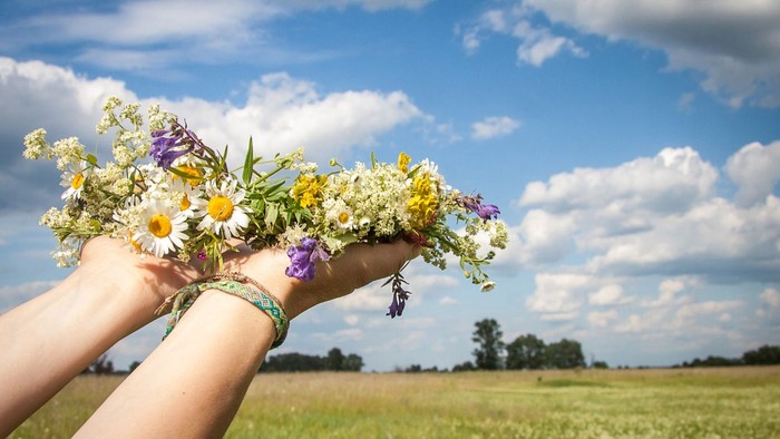 Hände, die einen leuchtenden Wildblumenkranz vor einem hellen, wolkenübersäten Himmel halten.