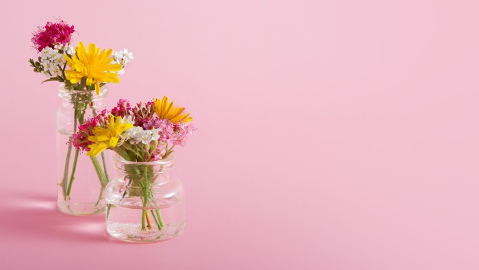 To klare vaser med livlige blomster sat op mod en blød lyserød baggrund.
