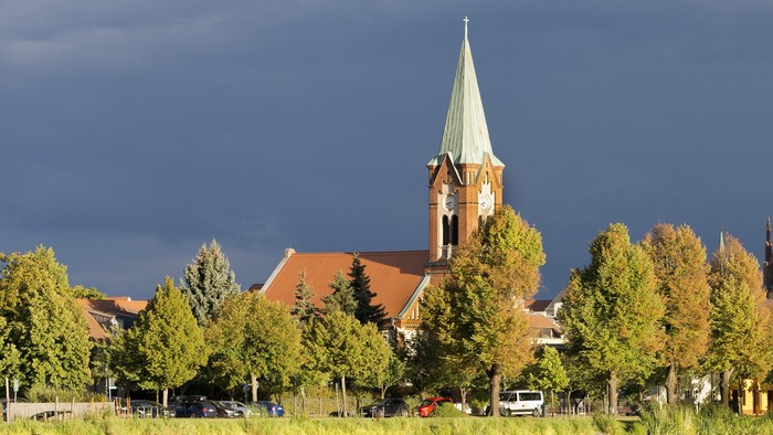 Eine beschauliche Kleinstadt am See mit historischen Kirchtürmen und üppigem Grün unter dramatischem, bewölktem Himmel.