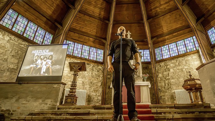 Ein Mann steht auf einer Bühne in einer Kirche und hält ein Mikrofon, während ein anderer Mann hinter ihm eine Gitarre spielt