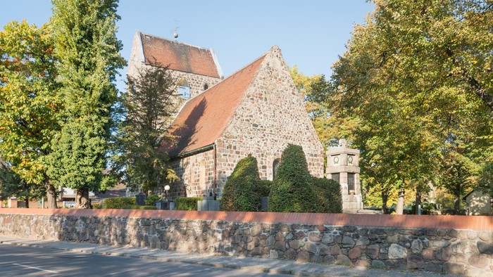 Das Bild zeigt eine Kirche mit einem roten Dach, umgeben von Bäumen und einem Steinzaun.