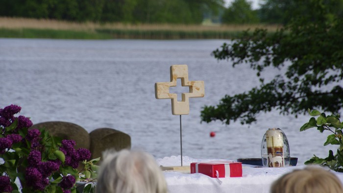 Eine Gruppe von Menschen versammelt sich am Wasser, mit einem Holzkreuz und einem Blumenstrauß.