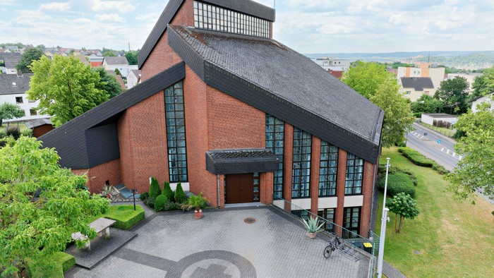 Das Bild zeigt eine moderne Kirche aus rotem Backstein mit großen Fenstern und einem gepflegten Vorhof.