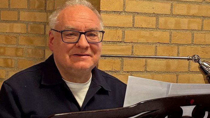 En mand med briller sidder ved et skrivebord med en åben bærbar computer og et orgelkeyboard i et hyggeligt rum.
