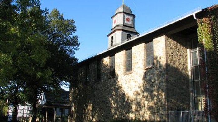 Steinerne Kirche mit hohem Turm und Kreuz, umgeben von Bäumen und Gras.