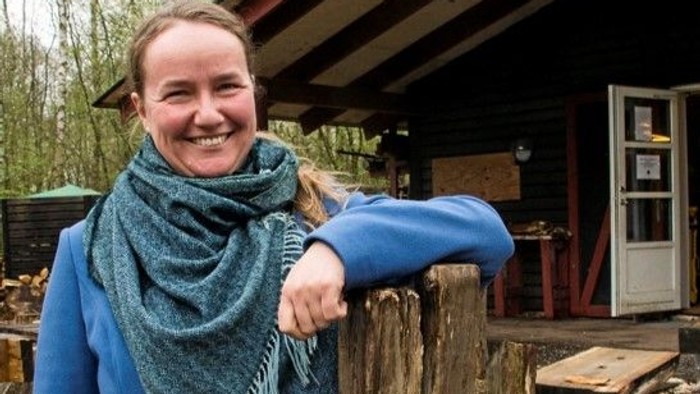 En kvinde i blå jakke og skarv smiler ved en træstamme i en have.