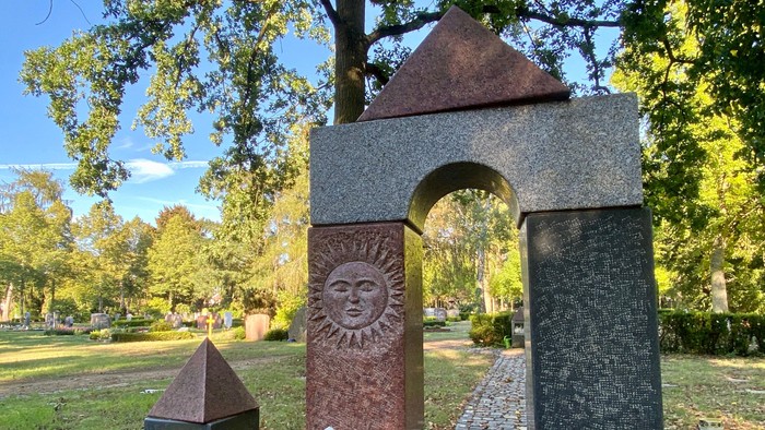 Modernes Grabdenkmal mit einem Sonnensymbol in einem Torbogen, umgeben von einem ruhigen Friedhof.