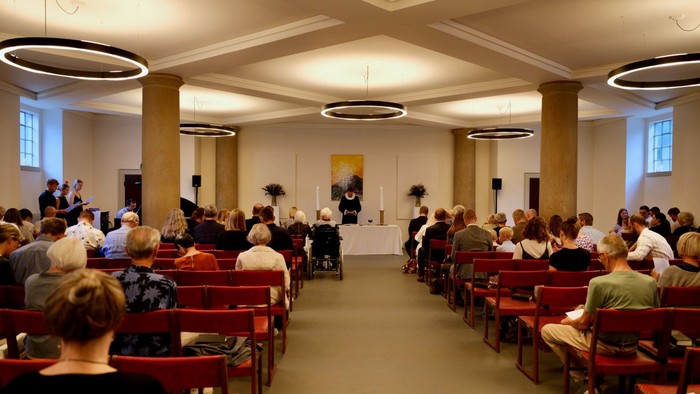 En stor sal med mange personer, der sidder på røde stolte og ser mod en scene med en person og et maleri.