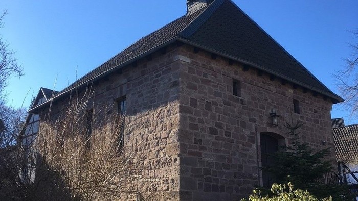 Steingebäude mit kleinem Turm und Wetterhahn, umgeben von Grünflächen unter einem klaren blauen Himmel.