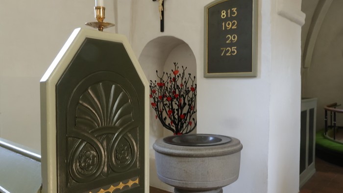 Elegant kirke døbefont med dekorativ plade, lysestage og blomsterarrangement ved siden af et kors.
