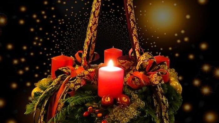 Ein Adventskranz mit drei Kerzen, geschmückt mit roten Bändern und grünen Blättern, hängt vor einem dunklen Hintergrund mit goldenen Sternen.