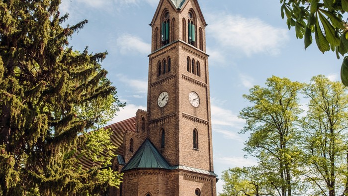 Kirche mit Uhrturm und grünem Dach, umgeben von Bäumen und Sträuchern.