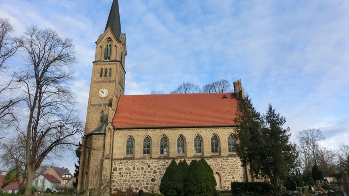 Kirche mit hohem Uhrturm und rotem Dach, umgeben von Bäumen und Sträuchern.