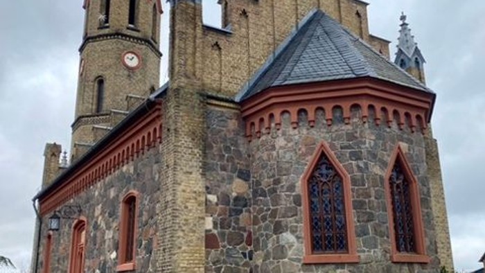 Steinerne Kirche mit roten Akzenten und hohem Turmspitz, umgeben von Grünflächen.