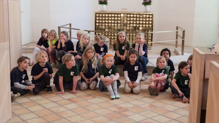 En gruppe børn sidder i en hal og lytter til en tale.