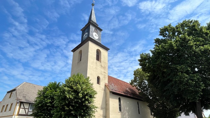 Kirchturm mit Uhr und Kreuz, umgeben von Bäumen und Häusern