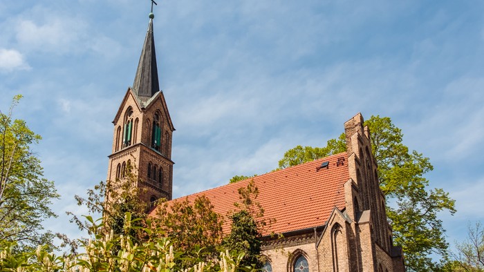 Kirche mit hohem Turmspitz und rotem Dach, umgeben von Bäumen unter blauem Himmel.