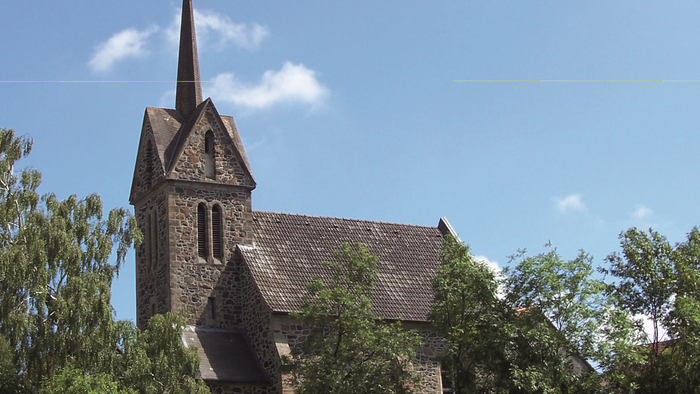 Kirchengebäude mit hohem Turm und spitzem Dach, umgeben von Bäumen unter blauem Himmel