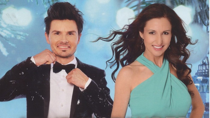 Zwei gut gekleidete Personen stehen vor einem blauen Hintergrund mit "ADVENTSZAUBER" beschriftet.