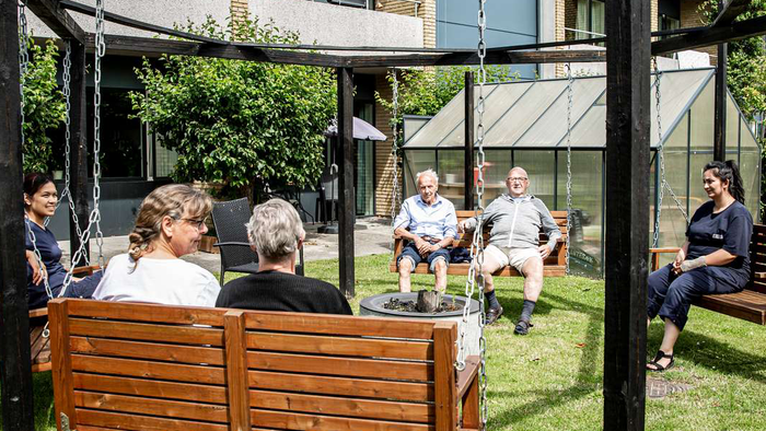 En gruppe mennesker sidder på bænke under en pergol i en have.