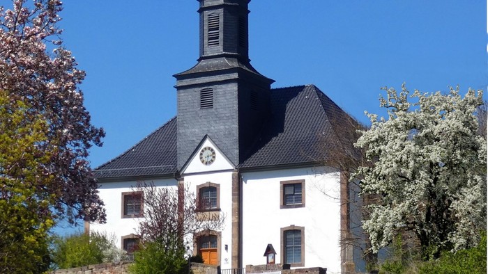 Weiße Kirche mit schwarzem Dach und hohem Turm, umgeben von Bäumen und Gras.