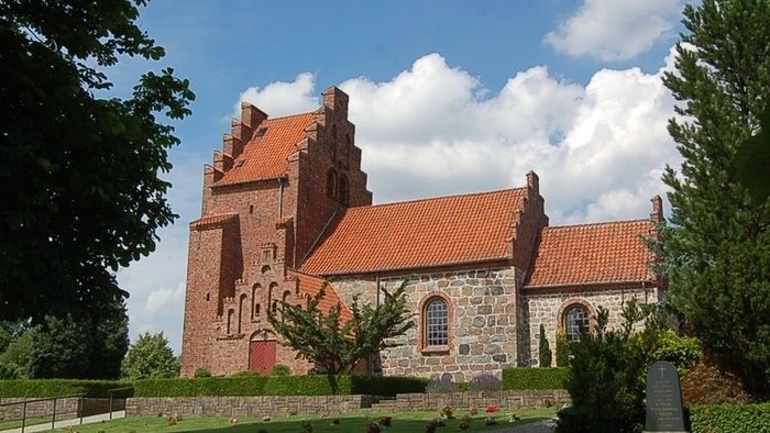 Gammel stenkirke med rødt tag og rød dør, omgivet af grøn græsplæne og træer.
