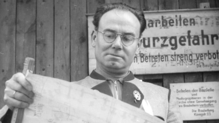 Mann mit Brille und Krawatte hält Zettel und Stift, vor Schildern mit deutscher Schrift.