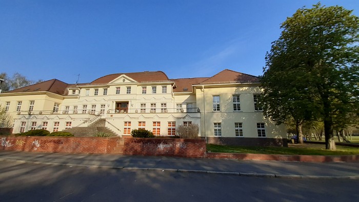 Großes, helles Gebäude mit rotem Dach und Ziegelmauer, Bäume und klarer blauer Himmel