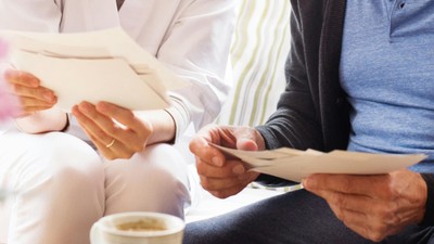 Zwei Personen sitzen an einem Tisch und lesen Papiere, eine Tasse Kaffee steht daneben.