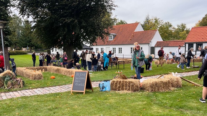 Udendørs arrangement med halmballer, folk der samles, og rustikke landsbyhuse i baggrunden.