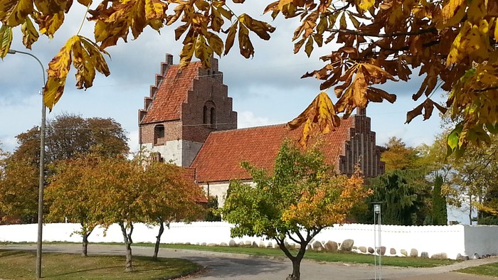 En kirke med røde tag og en mur, omgivet af faldne blade og træer