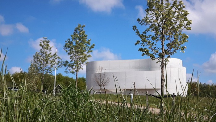 En moderne bygning med tre små træer foran, på en grøn eng under et blåt himmel.