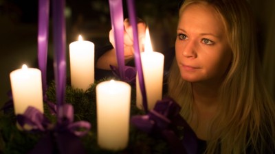 En ung pige med længe blond hår holder en adventskranse med lysende lygter og violet bænder.