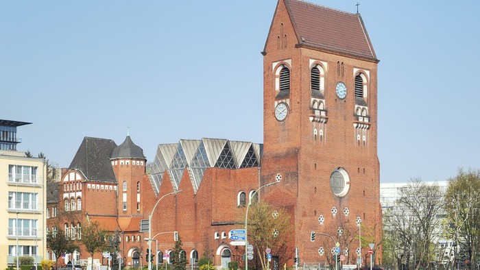 Historische Backsteinkirche mit hohem Uhrturm und modernen angrenzenden Gebäuden auf einem städtischen Platz.