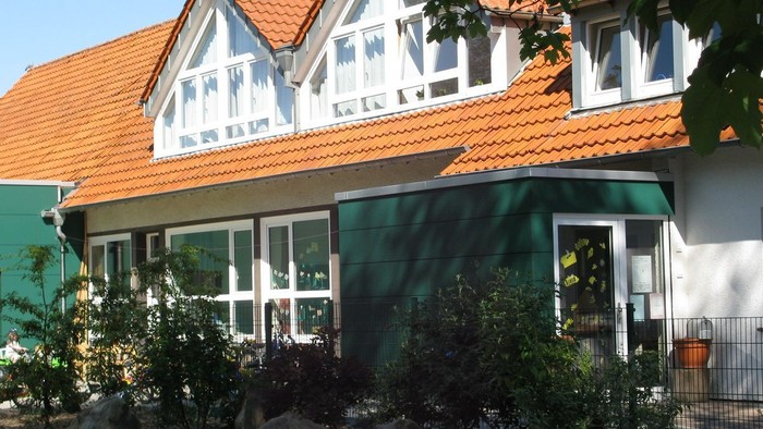 Das Bild zeigt ein charmantes Haus mit rotem Dach und grünen Fensterläden, umgeben von üppigem Grün.