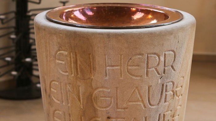 Der Taufstein ist aus Stein gefertigt und hat eine metallene Einfassung.