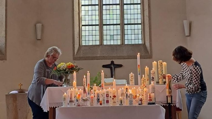 Zwei Personen zünden Kerzen an einem Gedenkaltar in einer ruhigen Kirche mit bunten Glasfenstern an.