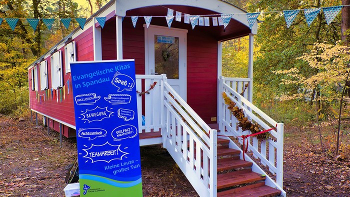 Ein rotes, kleines Gebäude mit weißer Treppe und einem blauen Banner davor, umgeben von Bäumen