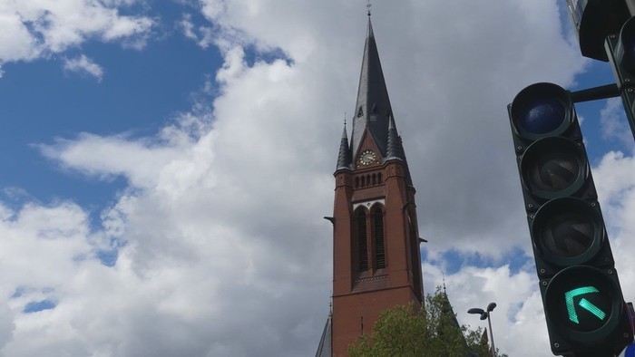 Ein roter Kirchturm mit spitzem Dach vor blauem Himmel und grünen Ampeln.
