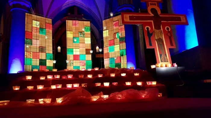 Beleuchteter Altarraum mit bunten Glasfenstern, vielen Kerzen auf Stufen und großem rotem Kreuz.