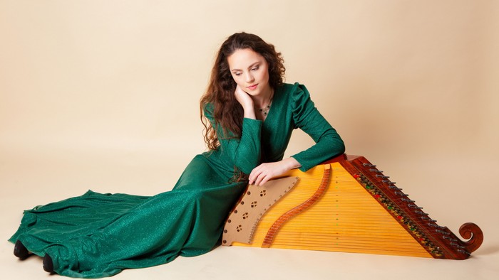A woman in a green dress plays a large, ornate yellow harp-like instrument thoughtfully.